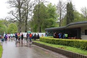 Evento di corsa in un parco sotto la pioggia, i partecipanti corrono o aspettano vicino a un edificio con punto ristoro.