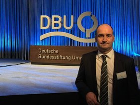Uomo in abito davanti a un palco con il logo della Fondazione Federale Tedesca per l’Ambiente (DBU) su sfondo illuminato di blu.